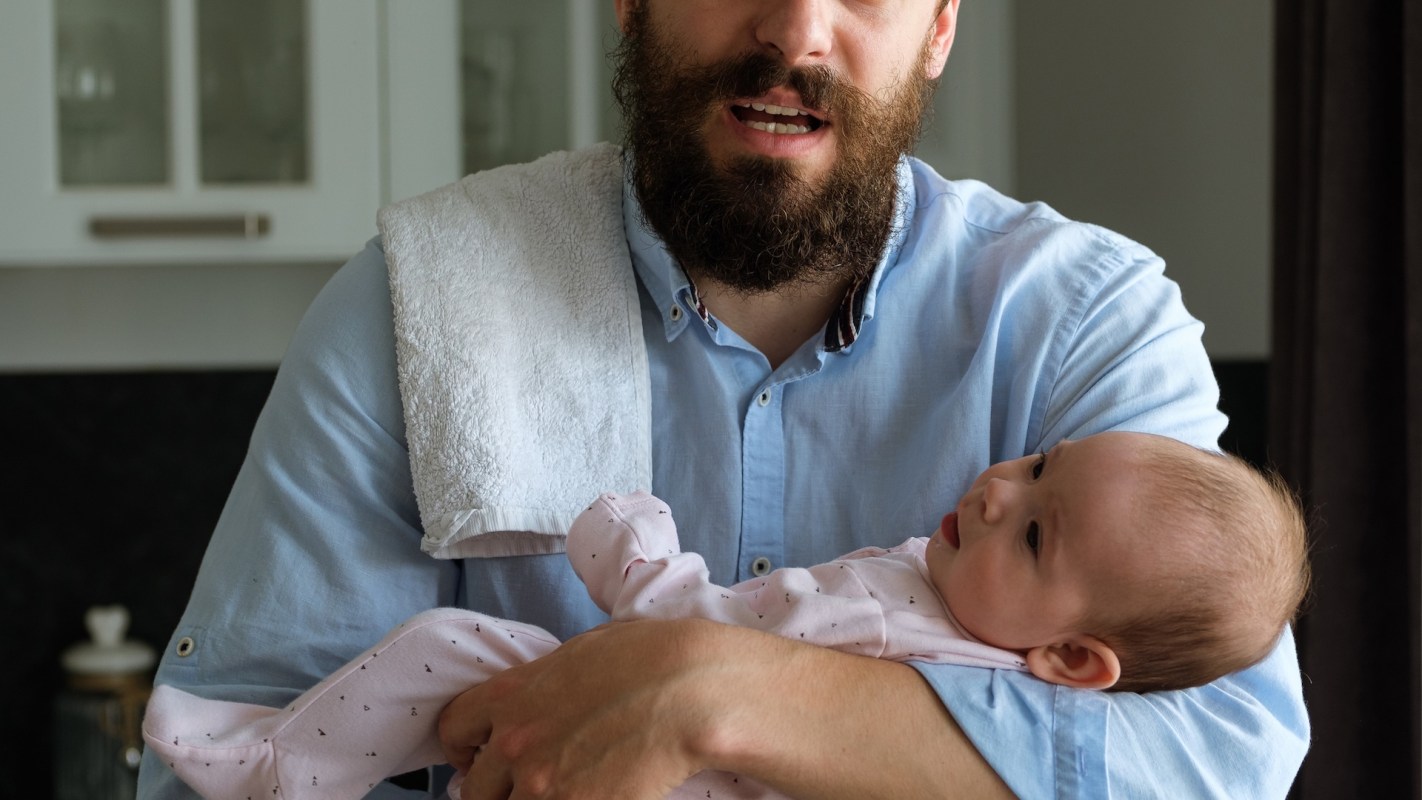 A bearded man in a blue shirt holds a baby dressed in pink, with a towel draped over his shoulder.
