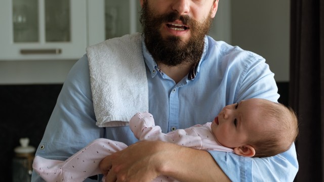 A bearded man in a blue shirt holds a baby dressed in pink, with a towel draped over his shoulder.