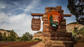 A sign for Zion National Park stands beside a scenic road with cliffs in the background.