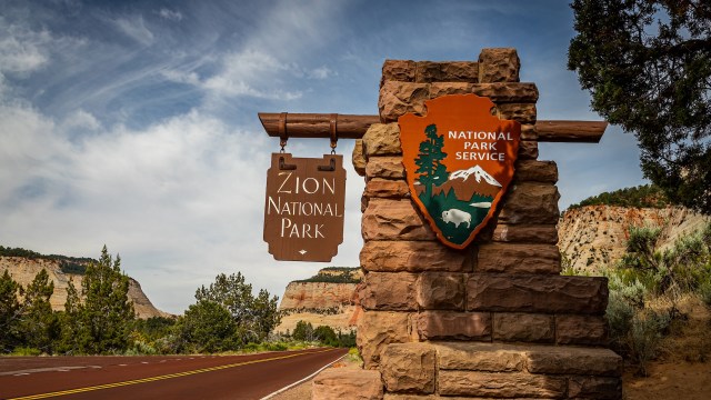 A sign for Zion National Park stands beside a scenic road with cliffs in the background.