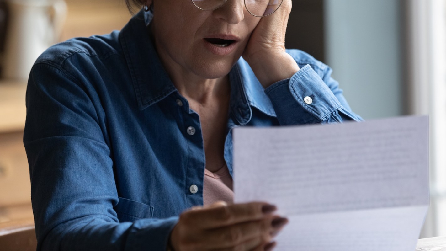 A senior woman with glasses looks shocked while looking at a paper document in her hand.