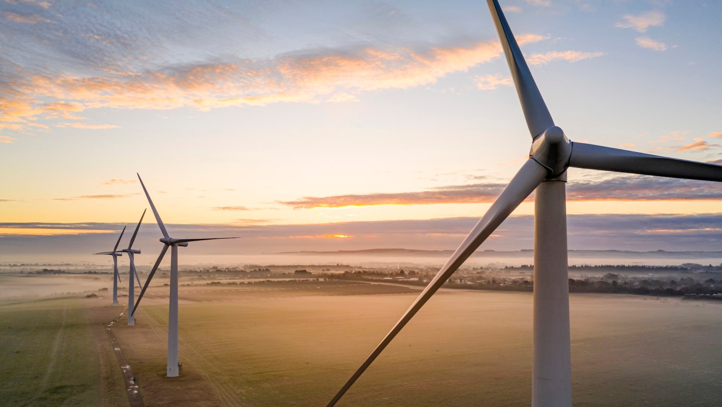 Wind turbines stand in a misty field, illuminated by a colorful sunrise against a cloudy sky.