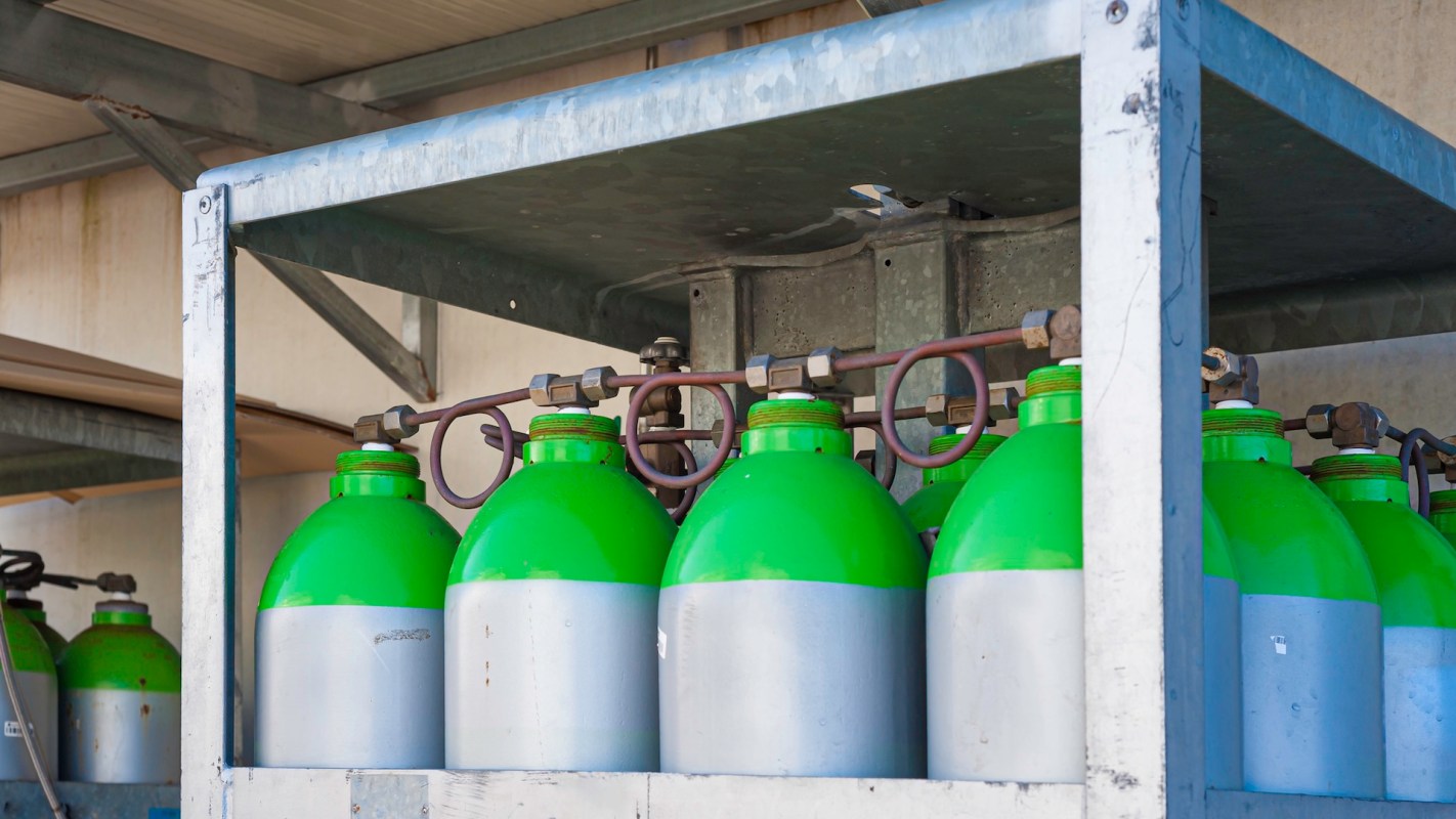 A rack of green and silver gas cylinders connected by pipes in a storage area.