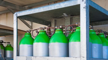 A rack of green and silver gas cylinders connected by pipes in a storage area.