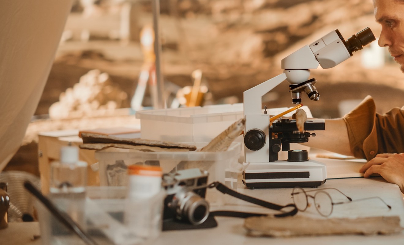 An archaeologist examines a specimen under a microscope on a cluttered research table at a dig site.