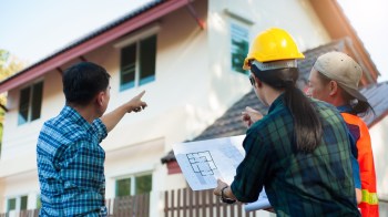 Three construction workers discuss plans while pointing at a building under construction in broad daylight.