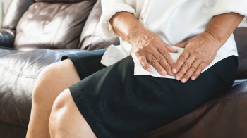 An older woman wearing a white shirt sits on a couch, hands rubbing her upper thigh.