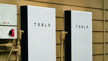 Two Tesla Powerwalls mounted on a brown exterior wall beside electrical components.