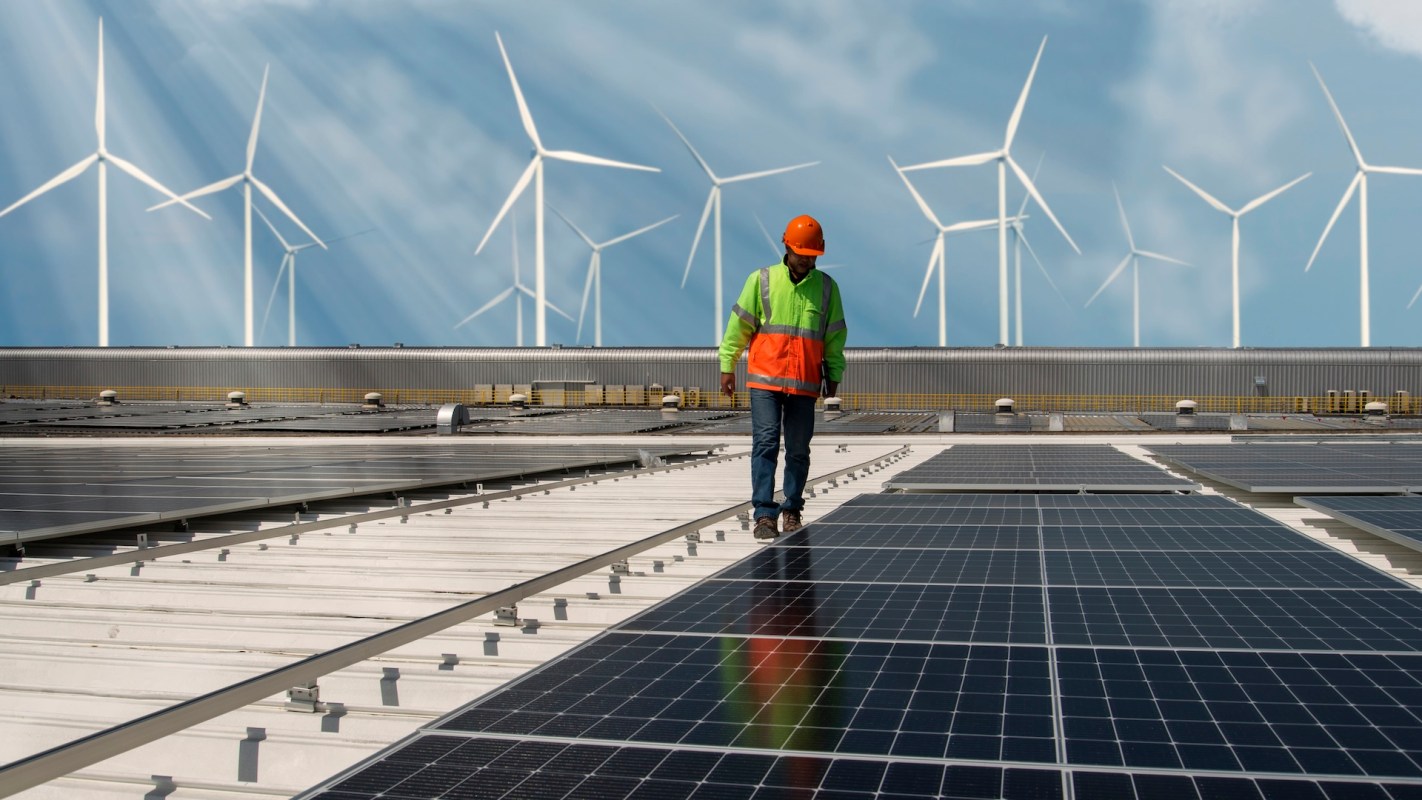 A worker in a safety vest walks on a rooftop covered in solar panels, with wind turbines visible in the background.