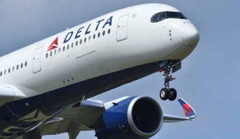 A Delta Airlines aircraft in flight with landing gear down against a blue sky.