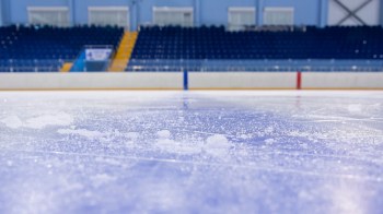 A close-up of an ice rink surface with scattered snow and blurred blue seats in the background.