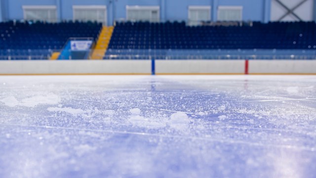 A close-up of an ice rink surface with scattered snow and blurred blue seats in the background.