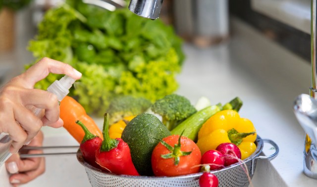 A hand sprays produce wash on vegetables in a colander.