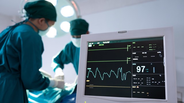 Surgeons in scrubs and masks work and monitor vital signs in an operating room.
