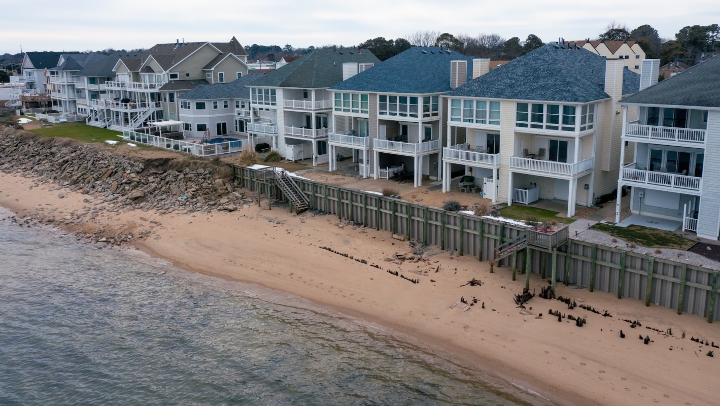 Aerial view of beachfront houses with sandy shore and protective wooden seawall along calm water.