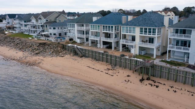 Aerial view of beachfront houses with sandy shore and protective wooden seawall along calm water.