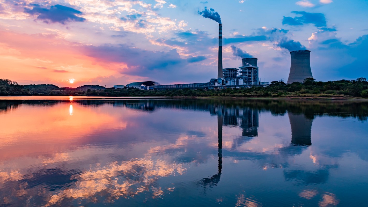 A nuclear power plant with cooling towers reflects in a calm lake at sunset, surrounded by colorful clouds.