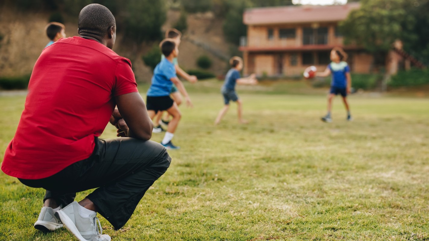 A person in a red shirt watches children playing soccer on a grassy field near a wooden building.