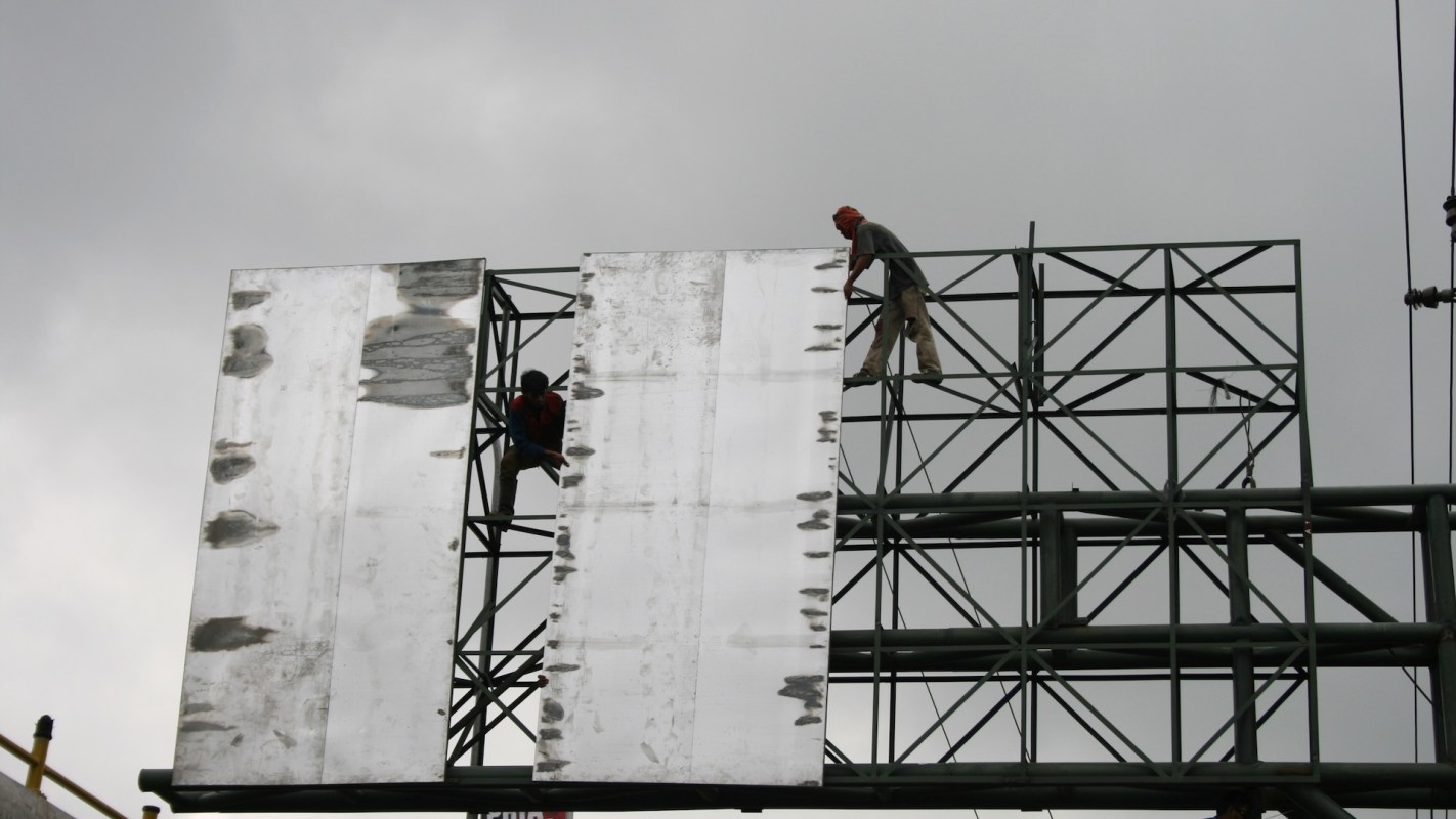 Two workers align large metal panels on a tall billboard frame against a cloudy sky.