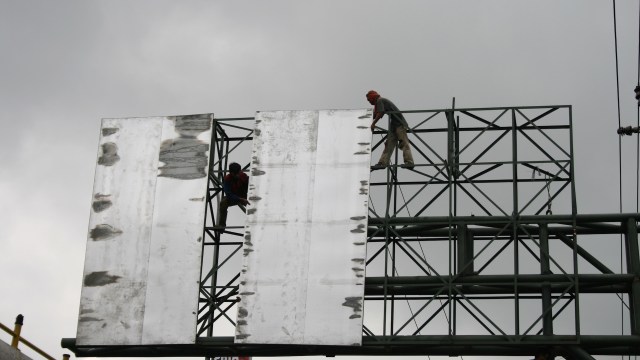 Two workers align large metal panels on a tall billboard frame against a cloudy sky.