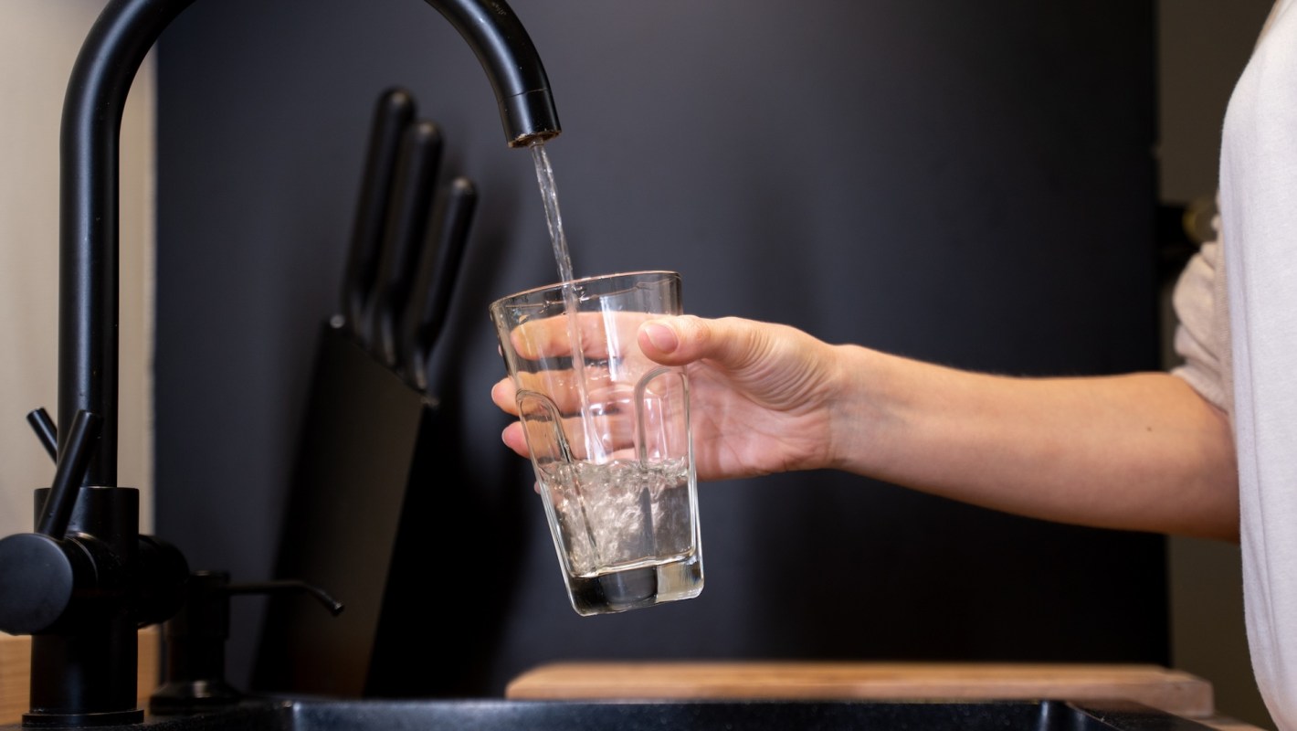 A person fills a clear glass with water from a modern black kitchen faucet.