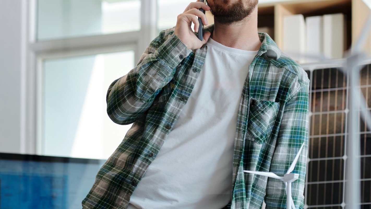A man in a plaid shirt talks on his phone in a modern office with a solar panel behind him and a model wind turbine nearby.