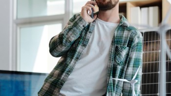 A man in a plaid shirt talks on his phone in a modern office with a solar panel behind him and a model wind turbine nearby.