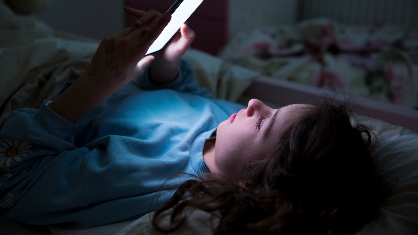 A child lies in bed, focused on a phone screen that illuminates their face in a dimly lit room.