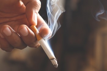 A close-up of a hand holding a lit cigarette, producing swirling smoke against a blurred background.