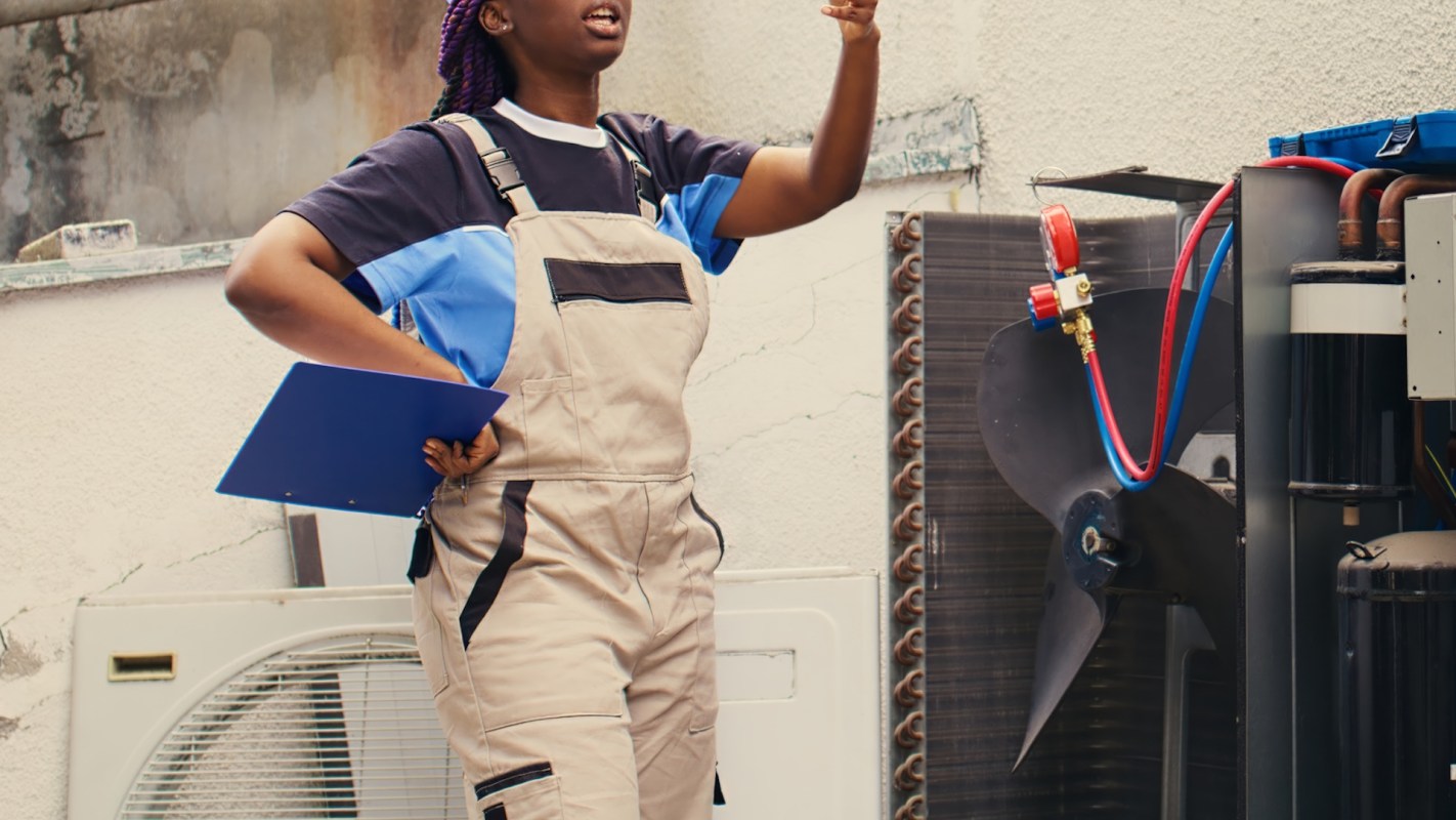 A woman in work overalls gestures while holding a clipboard near an air conditioning unit.