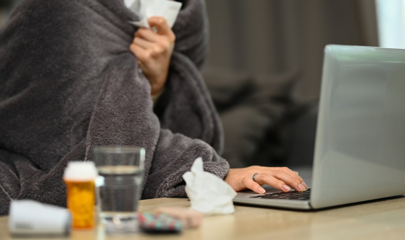 A person wrapped in a blanket sits at a table with a laptop, tissues, and medication.