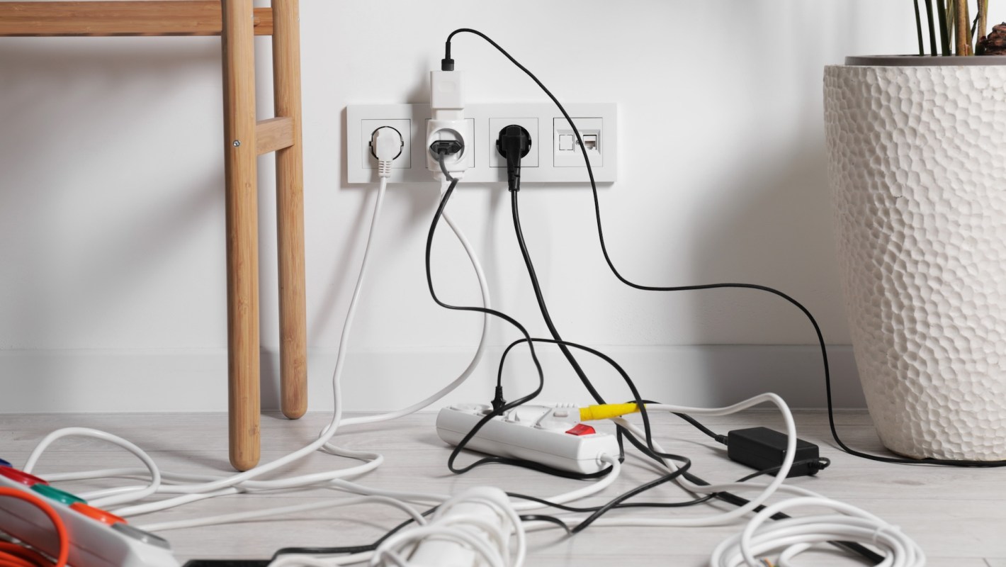 A jumble of electrical cords connected to a power strip on the floor next to a potted plant and a wooden table.