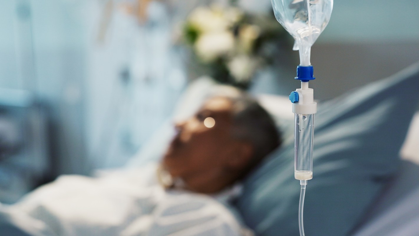 A person lies in a hospital bed, with an IV drip in the foreground.