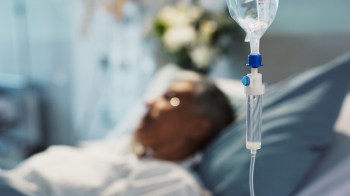 A person lies in a hospital bed, with an IV drip in the foreground.