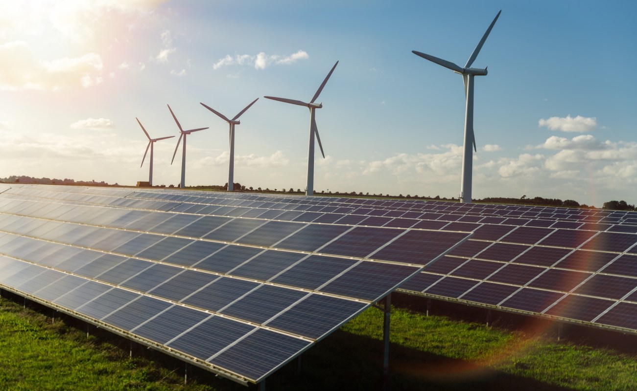 Solar panels in a field with wind turbines in the background under a clear blue sky.