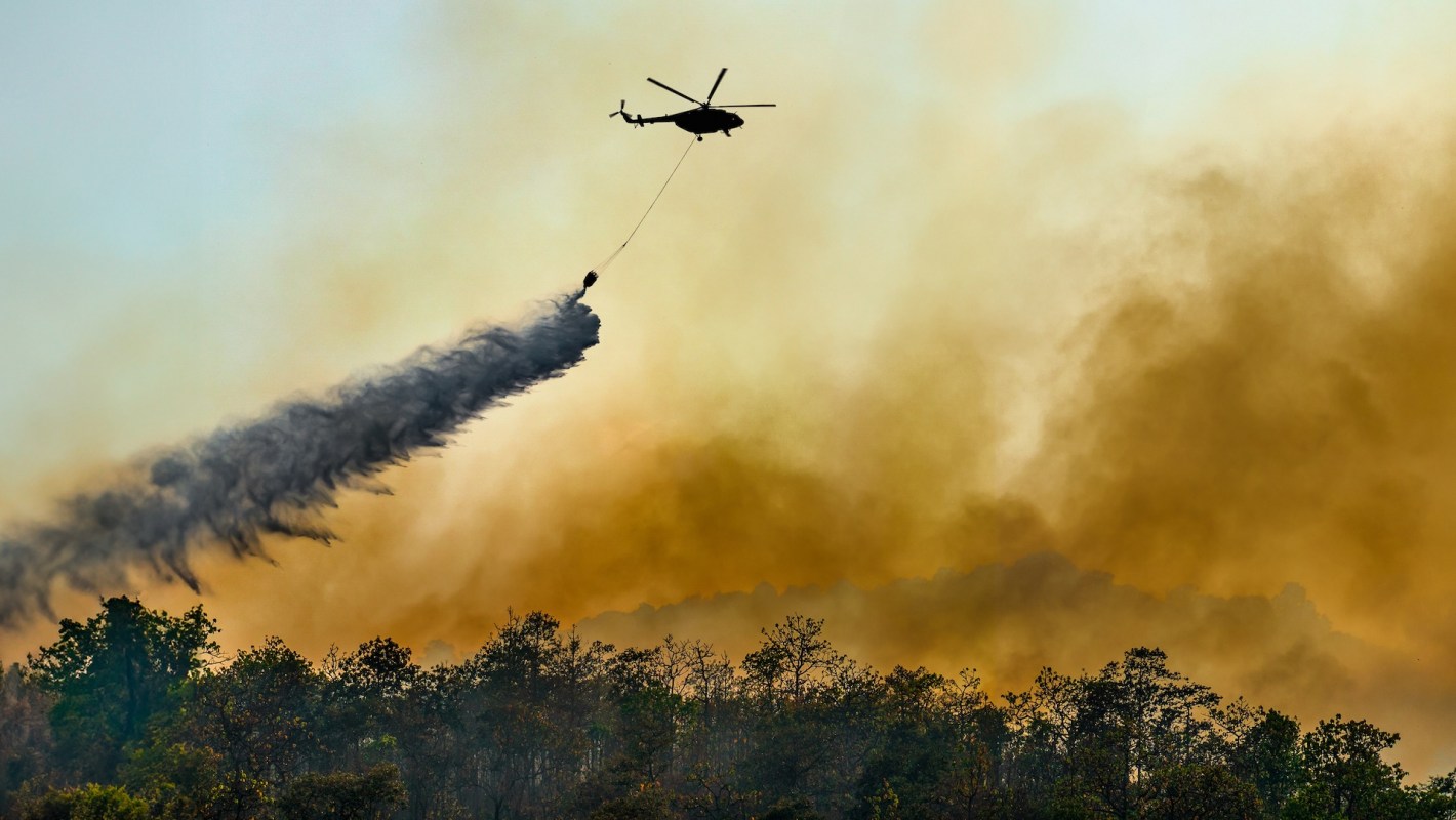 A helicopter drops water onto a forest fire, surrounded by billowing smoke and a hazy orange sky.