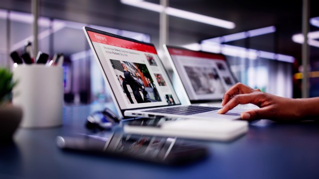 A person uses a laptop with news articles displayed, surrounded by office supplies and a smartphone.