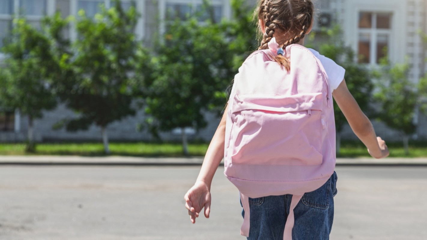 A girl with braids walks away, carrying a pink backpack in front of green trees and a building.