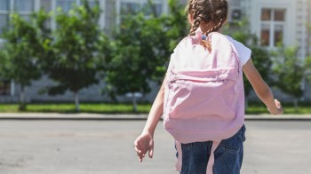 A girl with braids walks away, carrying a pink backpack in front of green trees and a building.