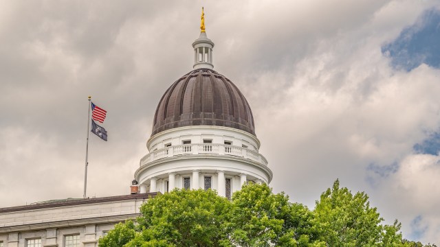 The dome of the Maine state capitol building is topped with a golden statue, surrounded by trees and flags against a cloudy sky.