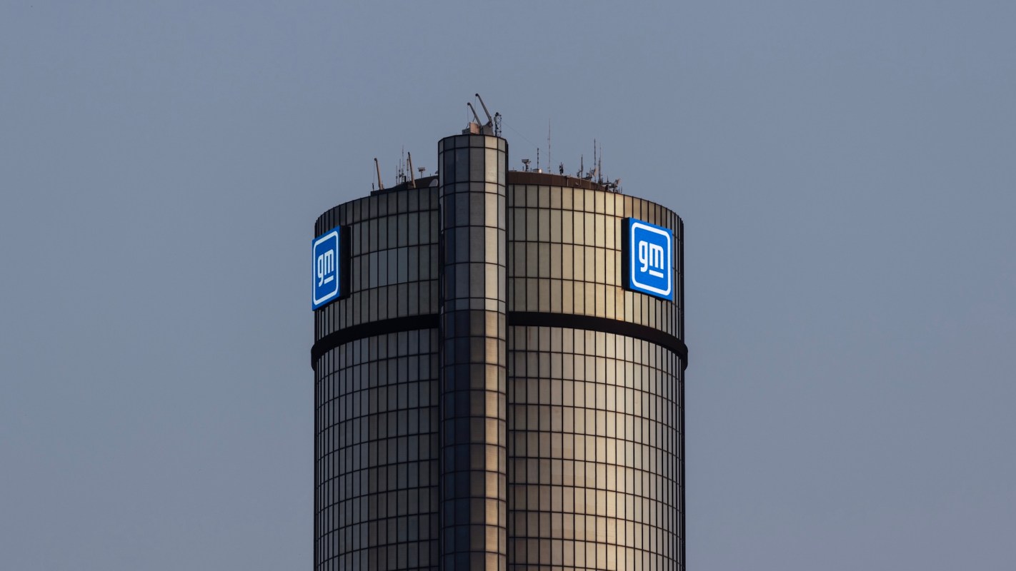 A close-up of the GM logo on top of a tall, modern glass building against a gray sky.