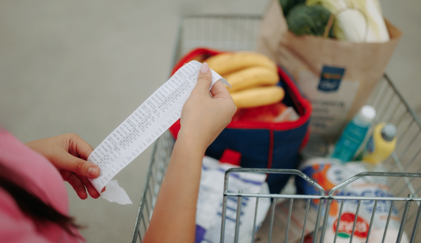 A person examines a grocery receipt while standing next to a shopping cart filled with various items.