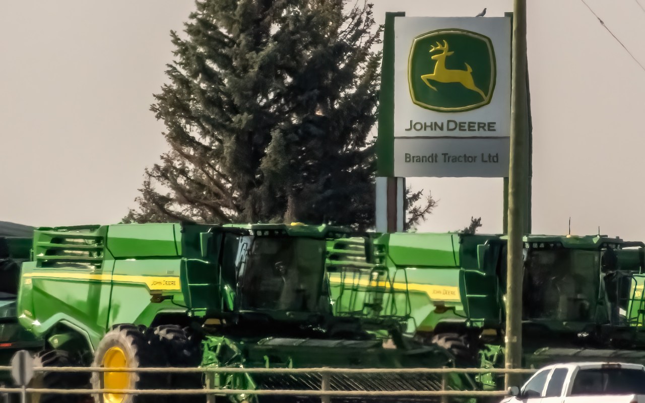 A row of green John Deere farming equipment beside a John Deere/Brandt Tractor sign and a tree in the background.