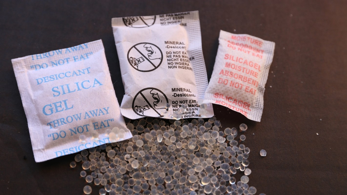 Three desiccant packets labeled "Silica Gel" and "Mineral Desiccant" are shown on a black surface with scattered gel beads.