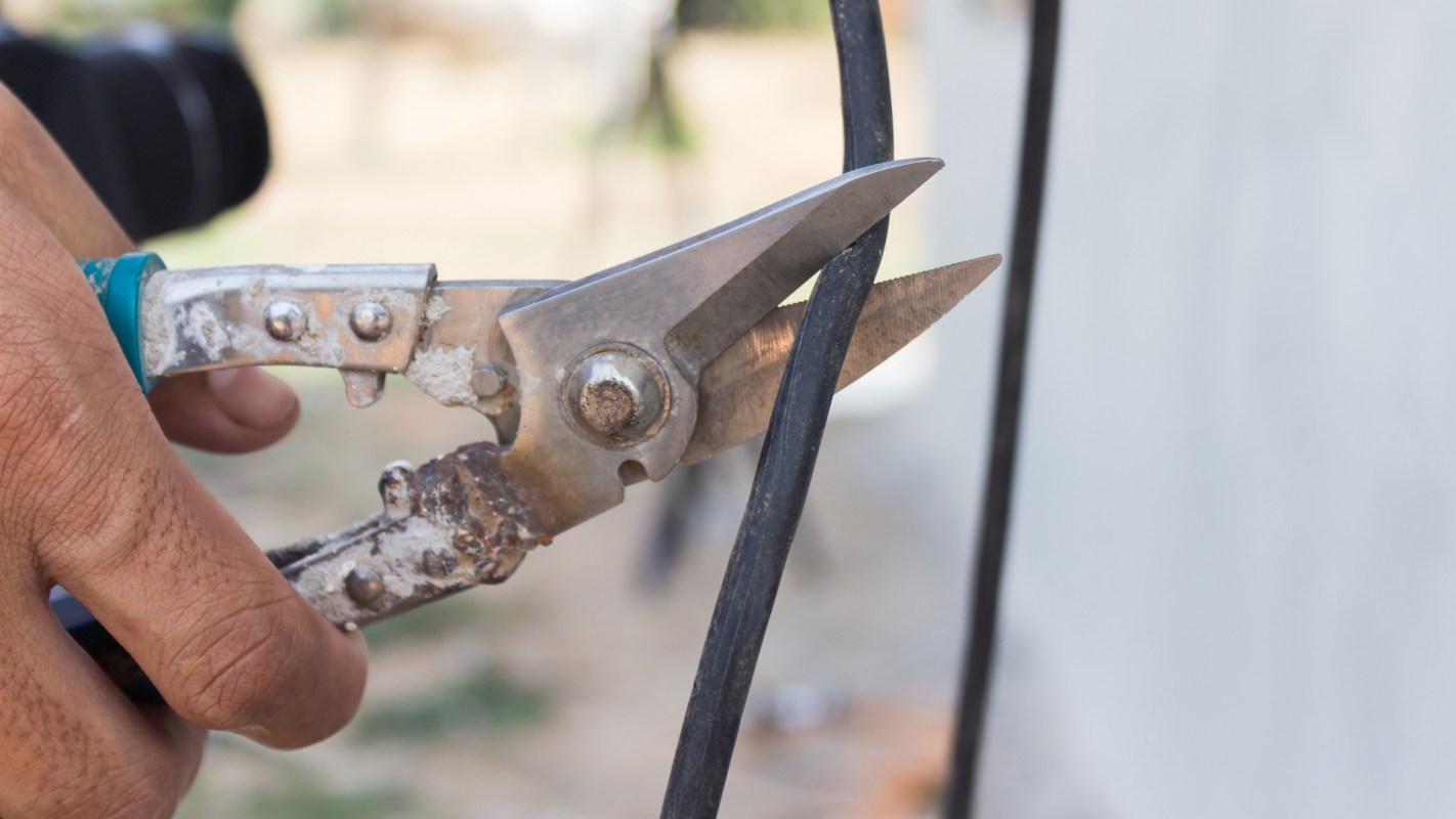 A person using a wirecutter to cut a thick black cable in an outdoor setting.