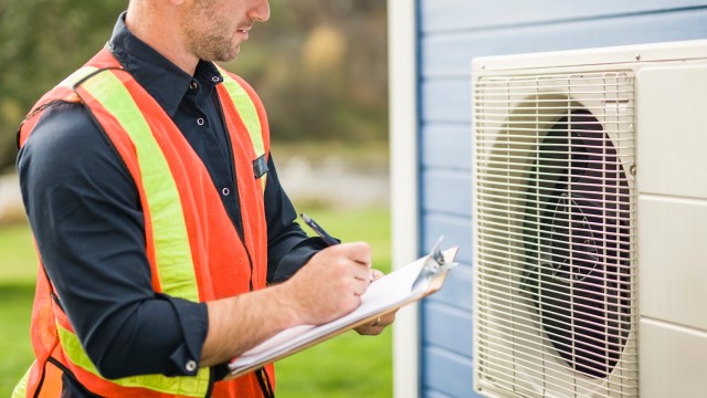 A worker in a safety vest inspects an air conditioning unit while taking notes on a clipboard.