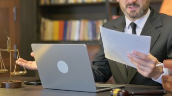 A suited man sits at a desk with a laptop, holding a document and gesturing in astonishment, with a scale of justice nearby.