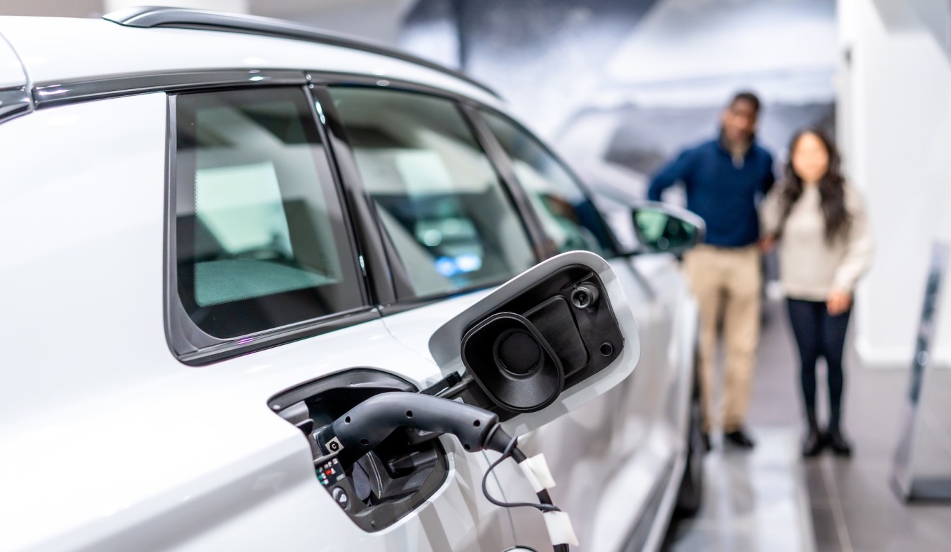 An electric vehicle's charging port is shown, with a couple in the background looking at it in a showroom.