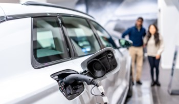 An electric vehicle's charging port is shown, with a couple in the background looking at it in a showroom.