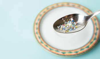 A spoonful of colorful microplastic fragments rests over a decorative plate against a light blue background.